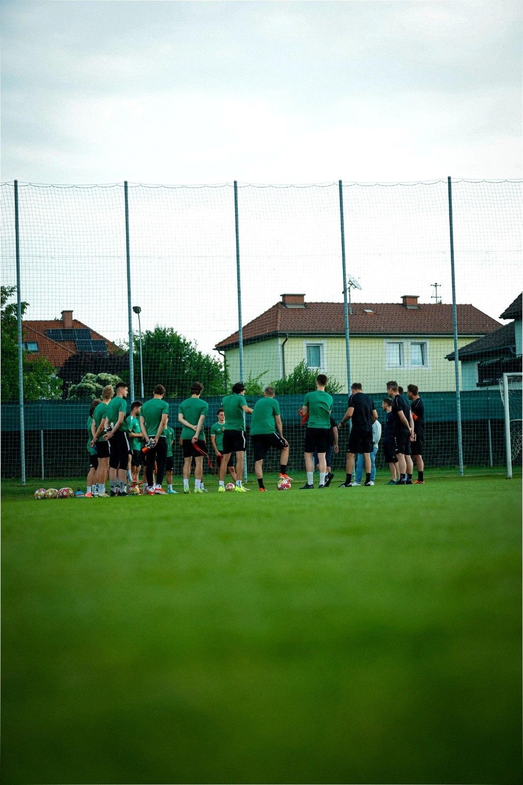 Eine Fußballmannschaft in grünen Uniformen trainiert auf einem Feld mit Netzzaun. Im Hintergrund sind Häuser zu sehen.