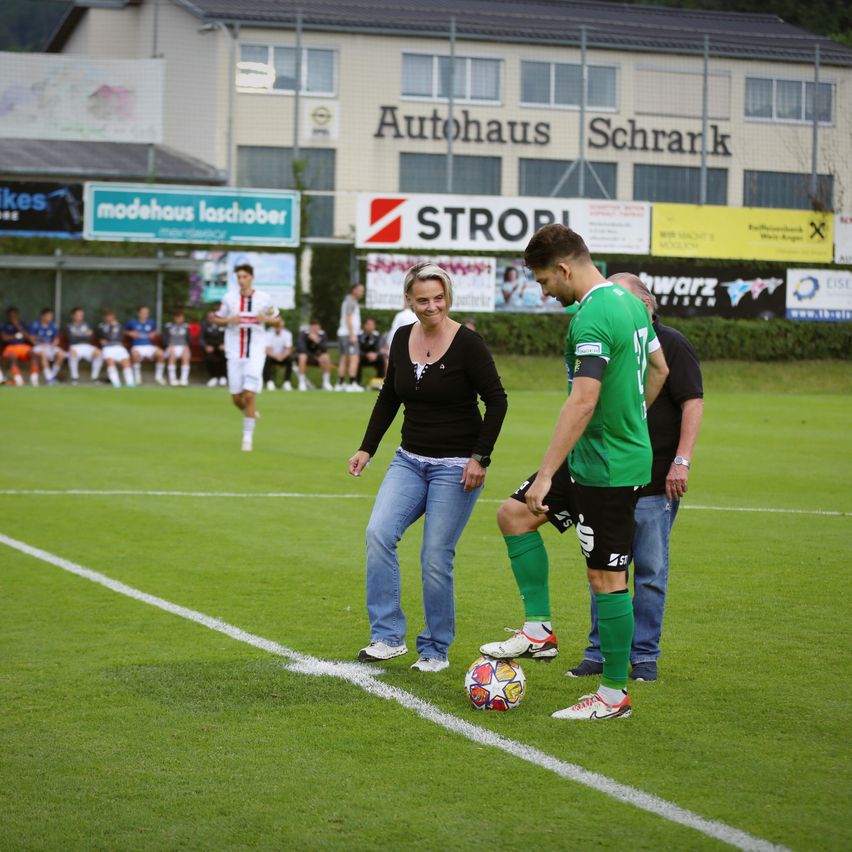Ein Fußballspieler nimmt einen Eckstoß mit einer Frau und einem Mann, die auf einem Feld zuschauen, mit einem Gebäude im Hintergrund.
