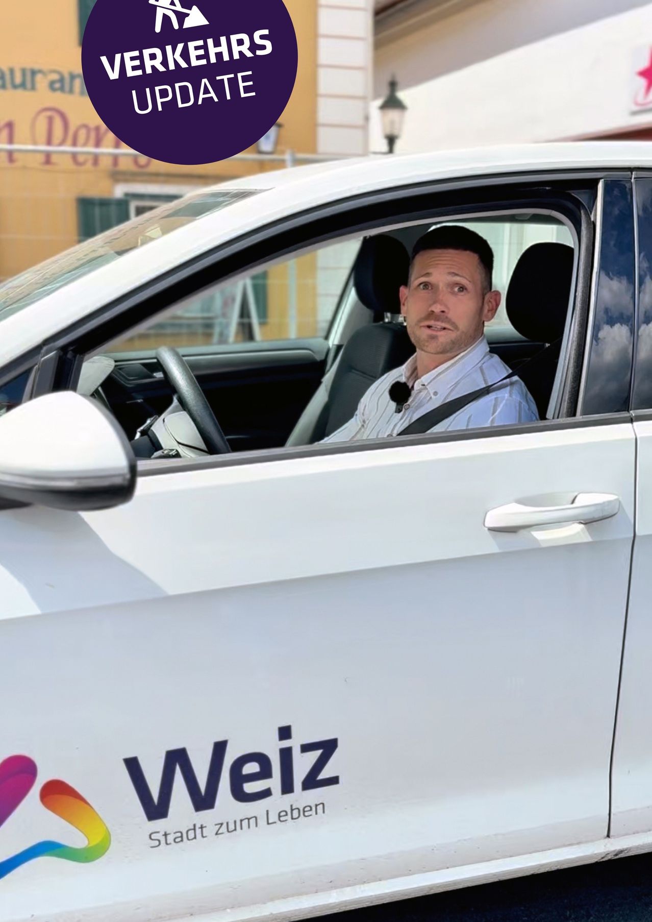 Ein Mann sitzt in einem weißen Weiz-Auto, trägt ein Mikrofon und ist vor einem Gebäude geparkt.