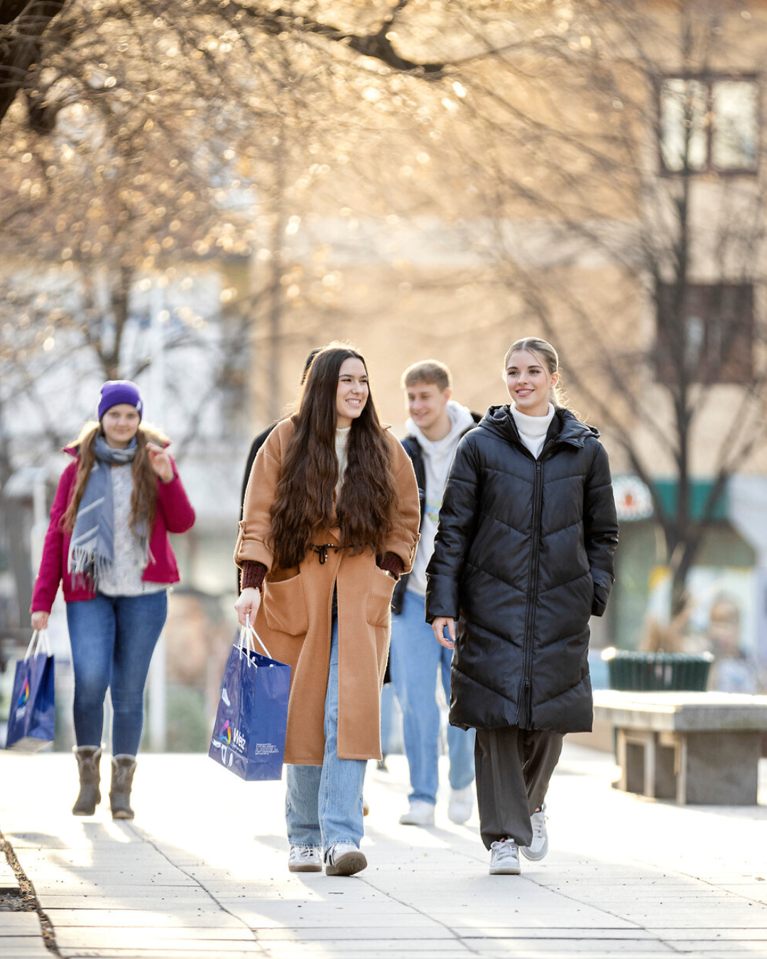 Eine Gruppe junger Menschen geht auf einer Stadtstraße. Zwei Frauen tragen Einkaufstüten. Eine trägt einen braunen Mantel, die andere eine schwarze Daunenjacke. Hinter ihnen trägt ein Mann einen weißen Hoodie. Bäume säumen den Bürgersteig.