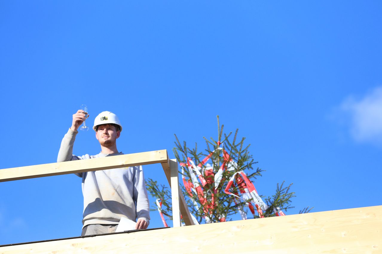 Ein Mann mit einem weißen Helm steht auf einer Holzplattform, hält ein Glas Wein und lächelt. Rechts steht ein geschmückter Weihnachtsbaum.