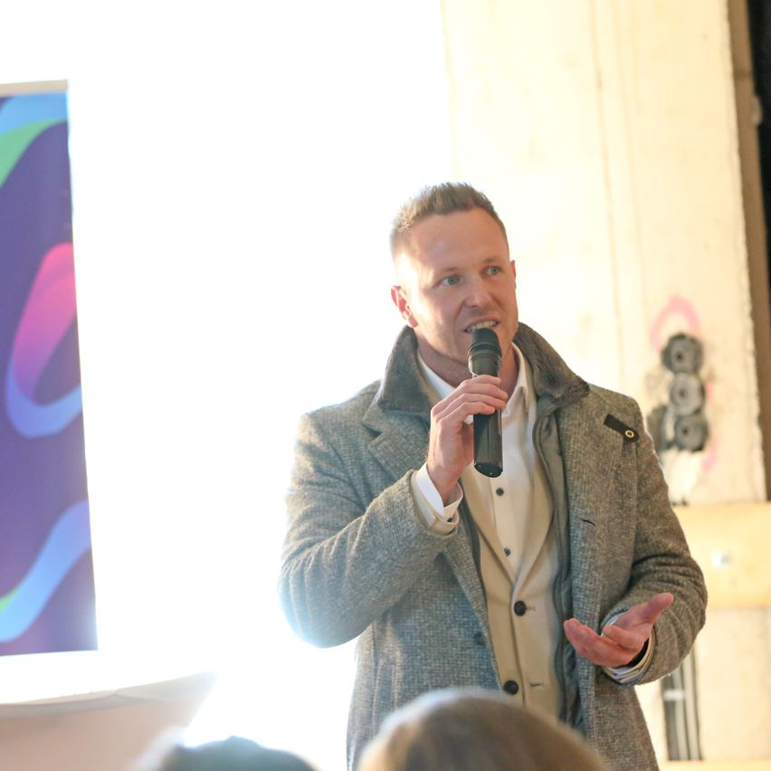 Ein Mann in einem grauen Mantel spricht in ein Mikrofon, während er ein Publikum anspricht, mit einem bunten Poster und einer Holzwand im Hintergrund.
