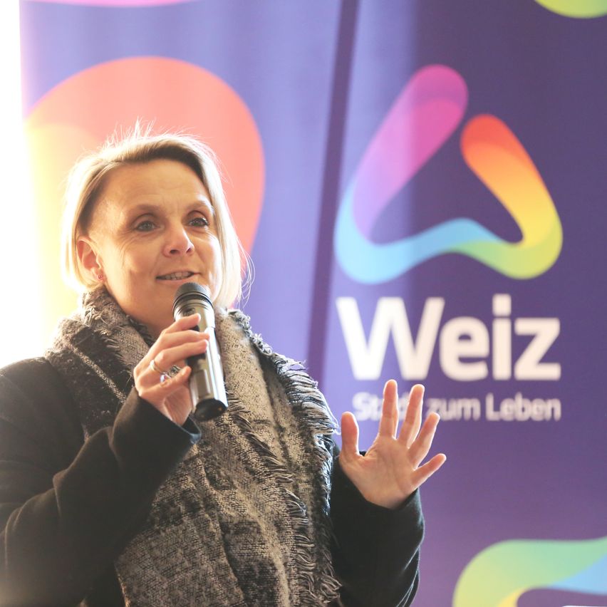 Eine Frau spricht in ein Mikrofon vor einem bunten Weiz-Banner. Sie trägt einen Schal und hat kurze blonde Haare.