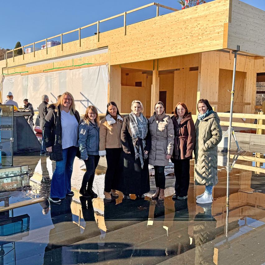 Sechs Frauen stehen vor einem teilweise gebauten Holzgebäude auf einer nassen Oberfläche mit sichtbaren Reflexionen. Hinter ihnen ist ein Mann anwesend.