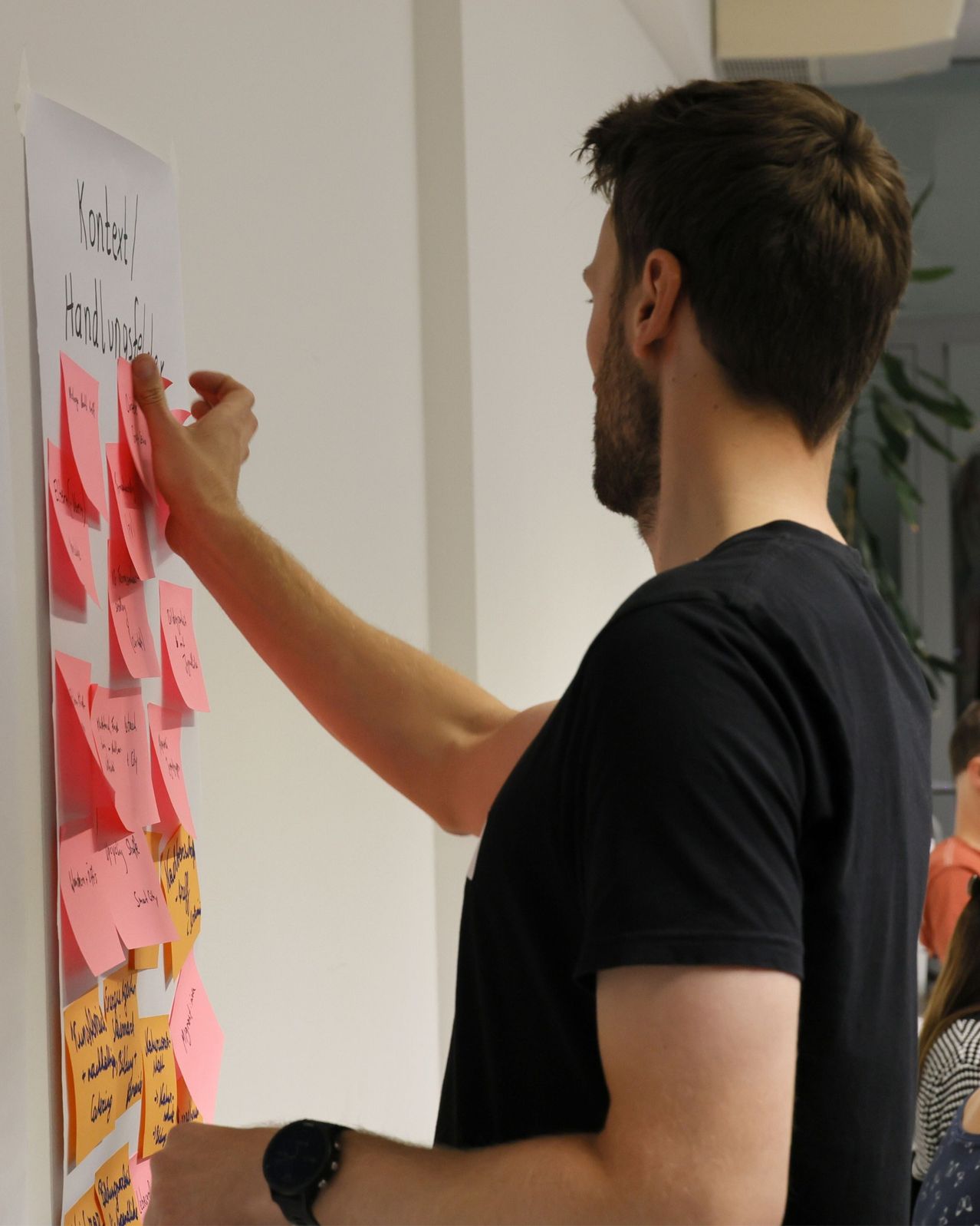Ein Mann fügt einen rosa Haftzettel an eine Wand, die bereits viele andere hat. Er trägt ein schwarzes T-Shirt.