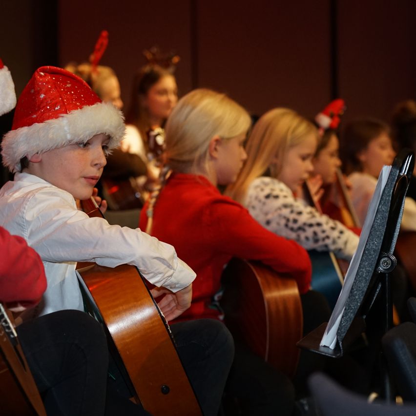 Mehrere Kinder in einem Chor, in Weihnachtsmützen gekleidet, spielen Gitarren und schauen auf einen Notenständer. Ein Junge mit einem Weihnachtsmannhut ist im Vordergrund.