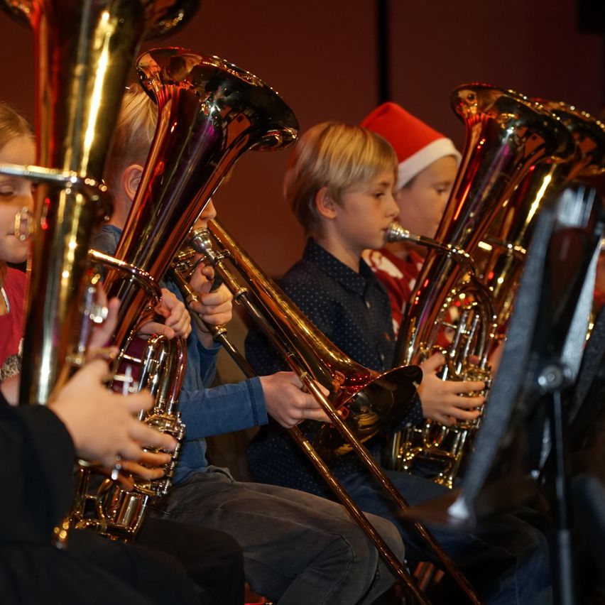 Eine Gruppe von Kindern spielt Posaunen in einer Band, wobei einer einen Weihnachtsmannhut trägt. Sie sitzen auf einer Bühne und führen wahrscheinlich bei einer musikalischen Veranstaltung auf.