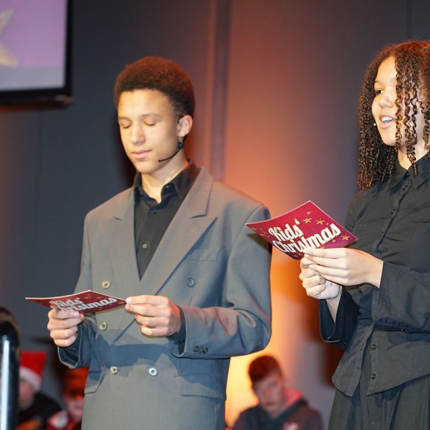 Zwei Personen auf der Bühne, eine hält ein rotes Büchlein mit dem Titel 'Kids Christmas' und die andere ein ähnliches Büchlein, beide schauen nach unten. Hinter ihnen ist ein verschwommener Hintergrund mit Menschen, die Weihnachtsmützen tragen.