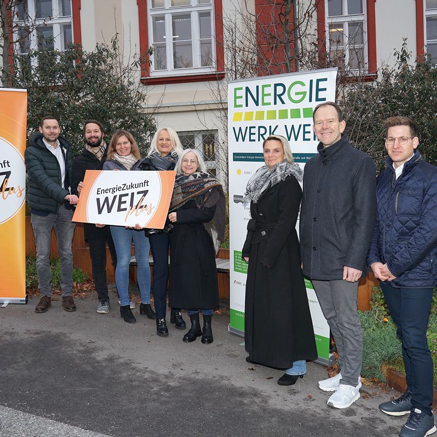 Eine Gruppe von Menschen steht vor einem Gebäude und hält ein Banner mit der Aufschrift 'EnergieZukunft WEIZ plus'. Sie tragen Winterkleidung und scheinen für ein Foto zu posieren.