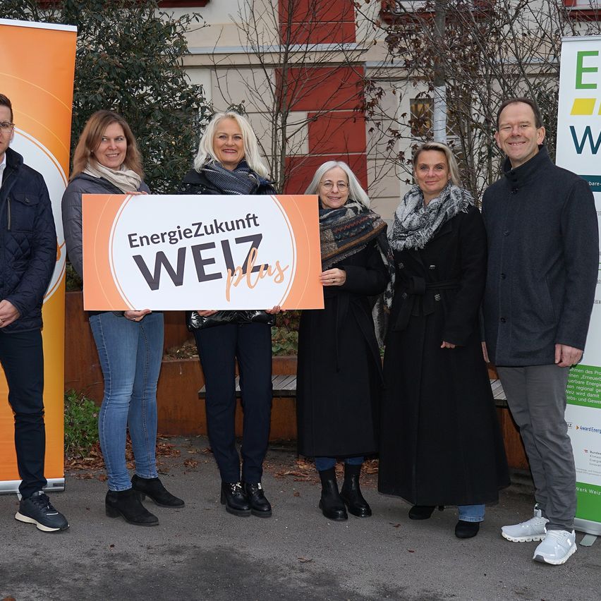 Fünf Personen stehen draußen und halten ein Schild mit der Aufschrift EnergieZukunft WEIZ plus. Sie alle lächeln. Im Hintergrund befinden sich Banner und ein Gebäude.