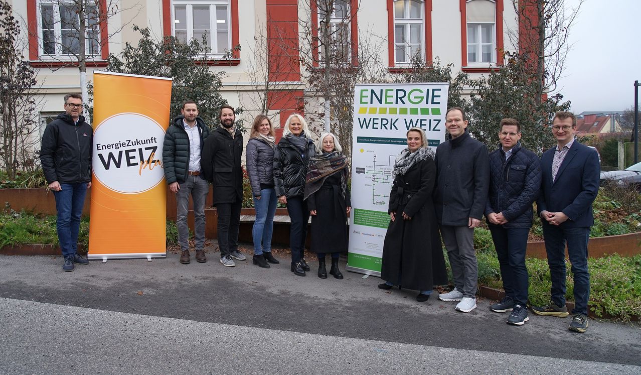 Eine Gruppe von Menschen steht draußen vor einem Gebäude und posiert für ein Foto. Sie tragen alle Winterkleidung. Ein Plakat ist hinter ihnen angebracht.