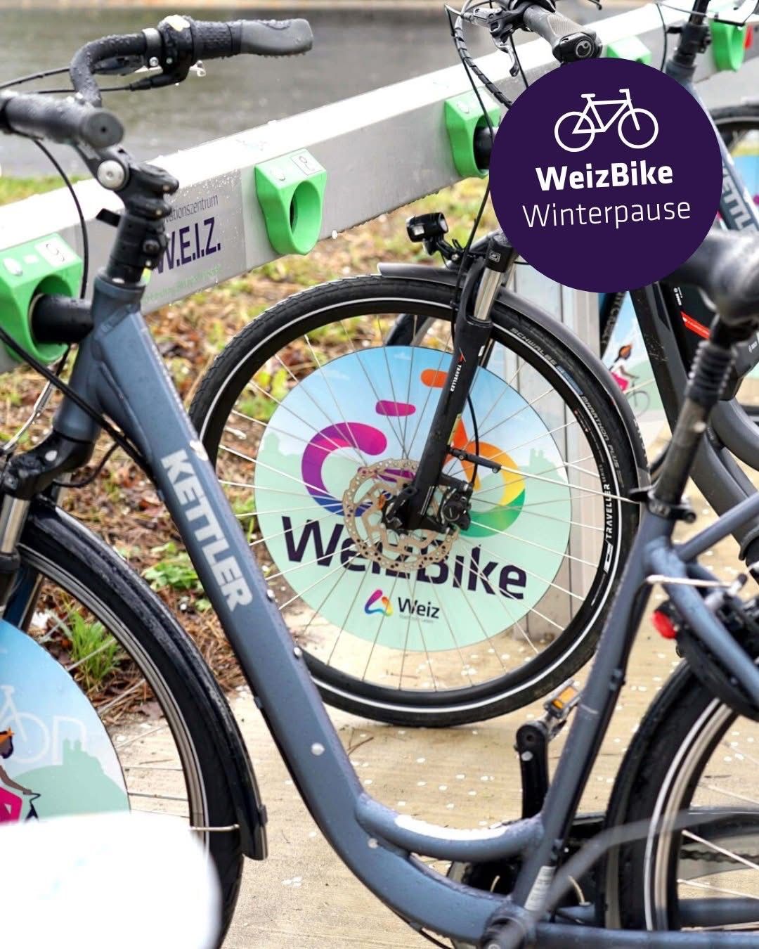 Mehrere Fahrräder in einer Reihe geparkt, eines davon mit einem farbenfrohen Rad. Es steht 'WeizBike' auf dem Rad und 'Winterpause' oben.