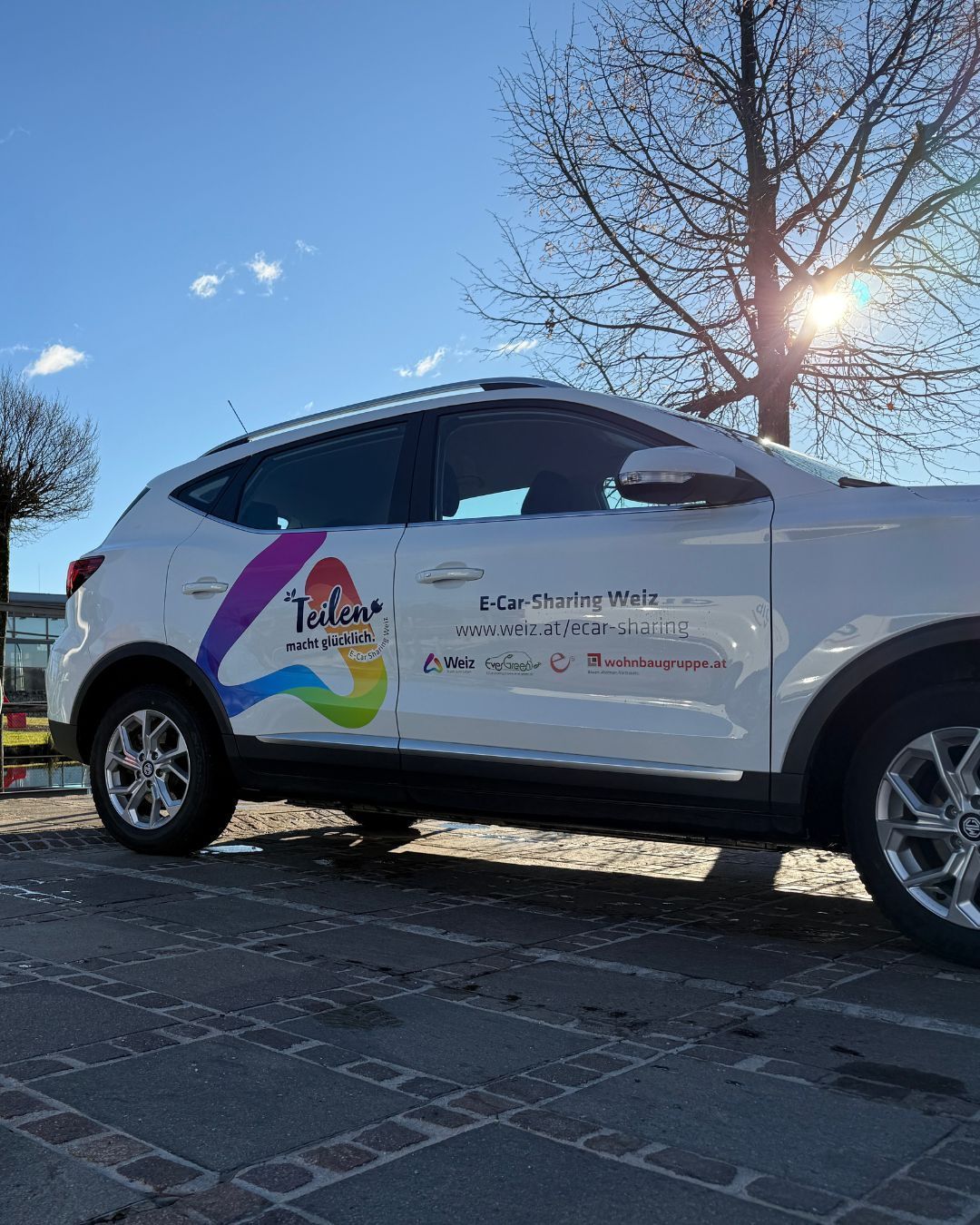 Ein weißer Elektrowagen steht auf einer gepflasterten Straße und zeigt das Logo und die Website von E-Car-Sharing Weiz.