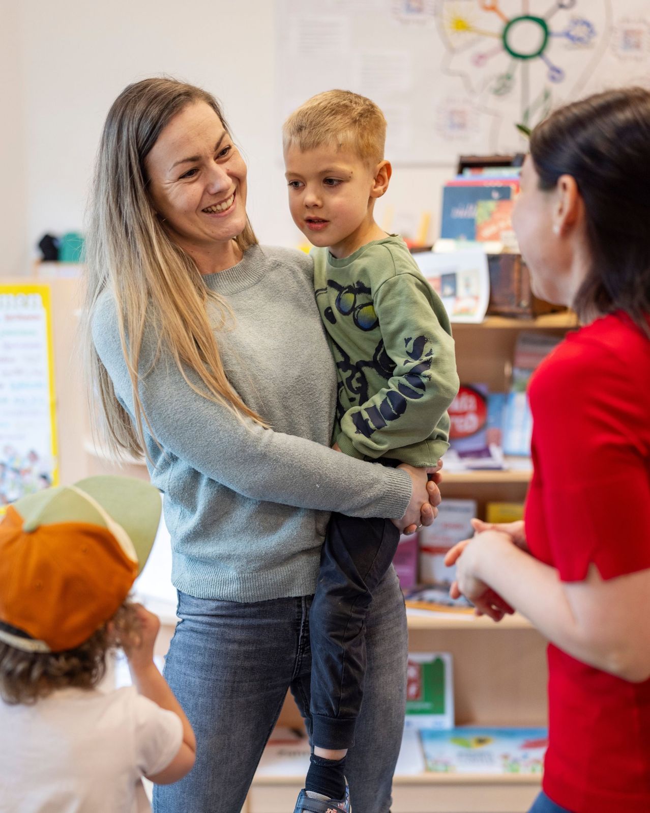 Eine Frau, die einen kleinen Jungen hält, lächelt eine Frau in Rot an. Hinter ihnen beobachtet ein Kind mit Hut. Sie befinden sich in einem Raum mit Bücherregalen.