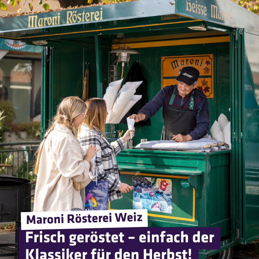 Ein Außenimbisswagen mit der Aufschrift Maroni Roesterei serviert frisch geröstete Kastanien. Zwei Frauen stehen vor dem Wagen. Ein Mann in schwarzer Kappe und Schürze bedient die Frauen.