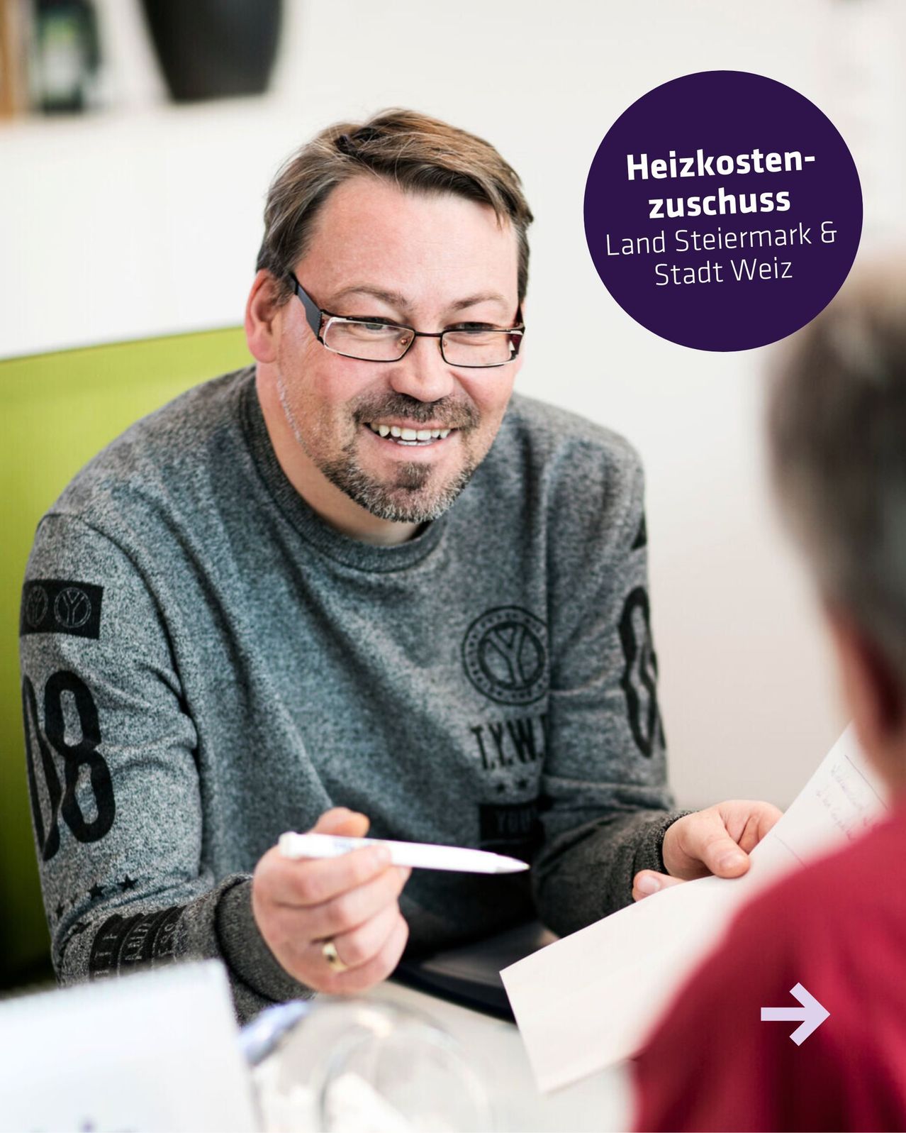 Ein Mann in einem grauen Sweatshirt sitzt an einem Schreibtisch, hält einen Stift und Papier in der Hand und lächelt eine andere Person an. Die Umgebung scheint ein Büro zu sein mit einem grünen Stuhl und einer weißen Wand.