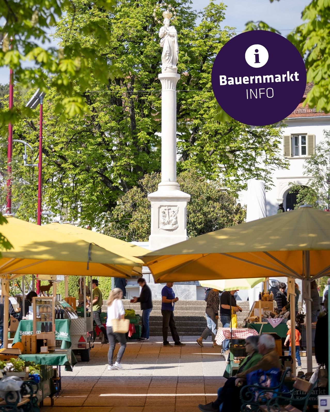 A farmers market with yellow umbrellas and a statue in the background. There are people shopping and a cart with bottles. Trees and a building are visible.