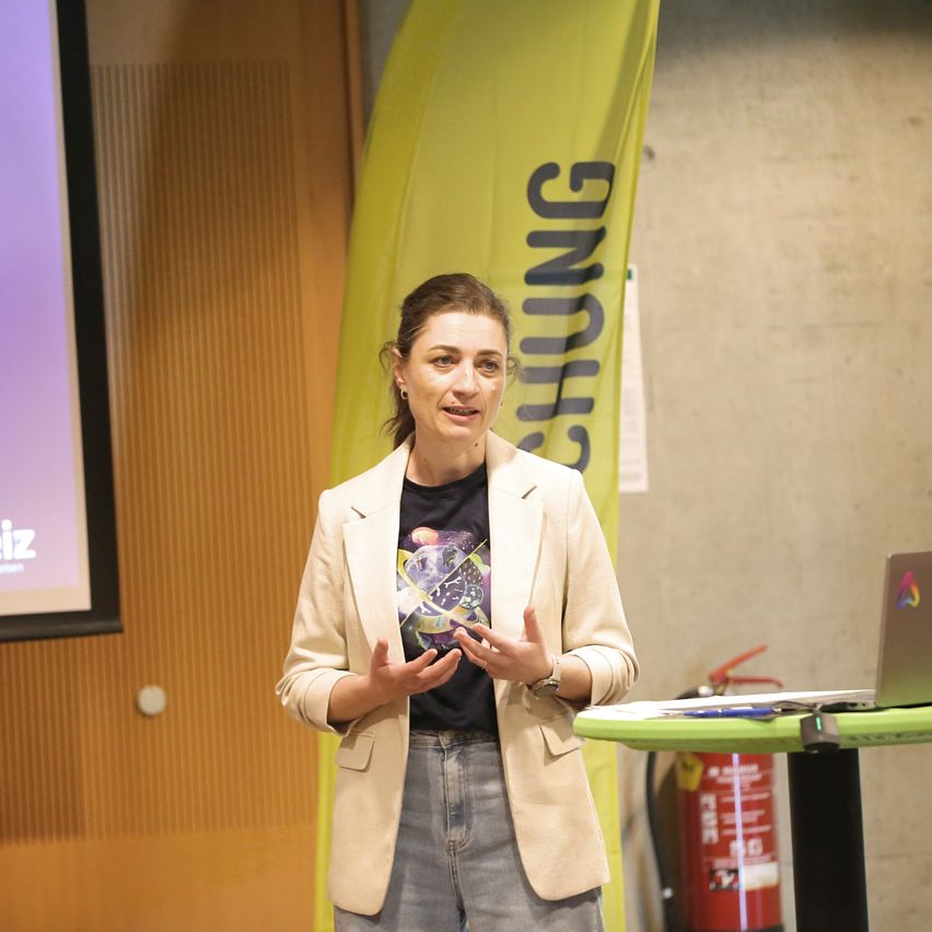 A woman stands in front of a screen giving a presentation. She has a light-colored blazer and a dark t-shirt. Behind her is a yellow banner with the word 'Gunning' on it.