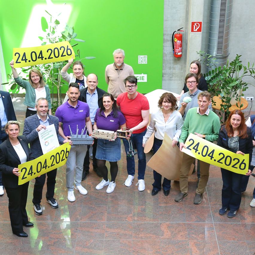 A group of people holding yellow signs with the date 24.04.2026. They are standing in a room with green walls and potted plants.