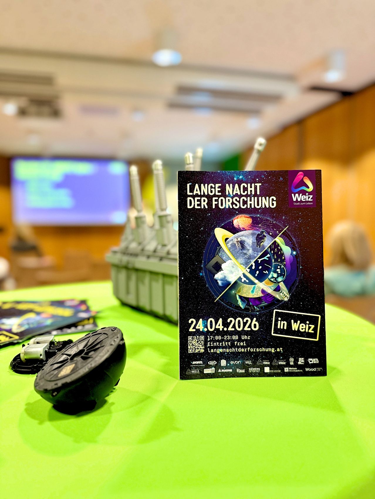 A poster titled Lange Nacht der Forschung in Weiz on a green table with a black object. Date 24.04.2026. Behind the table, a projector screen is visible.