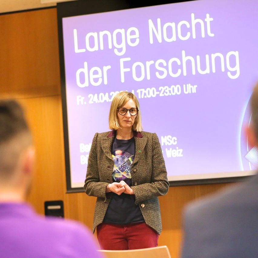 A woman stands in front of a presentation screen with the text 'Long Night of Research', holding her hands together.