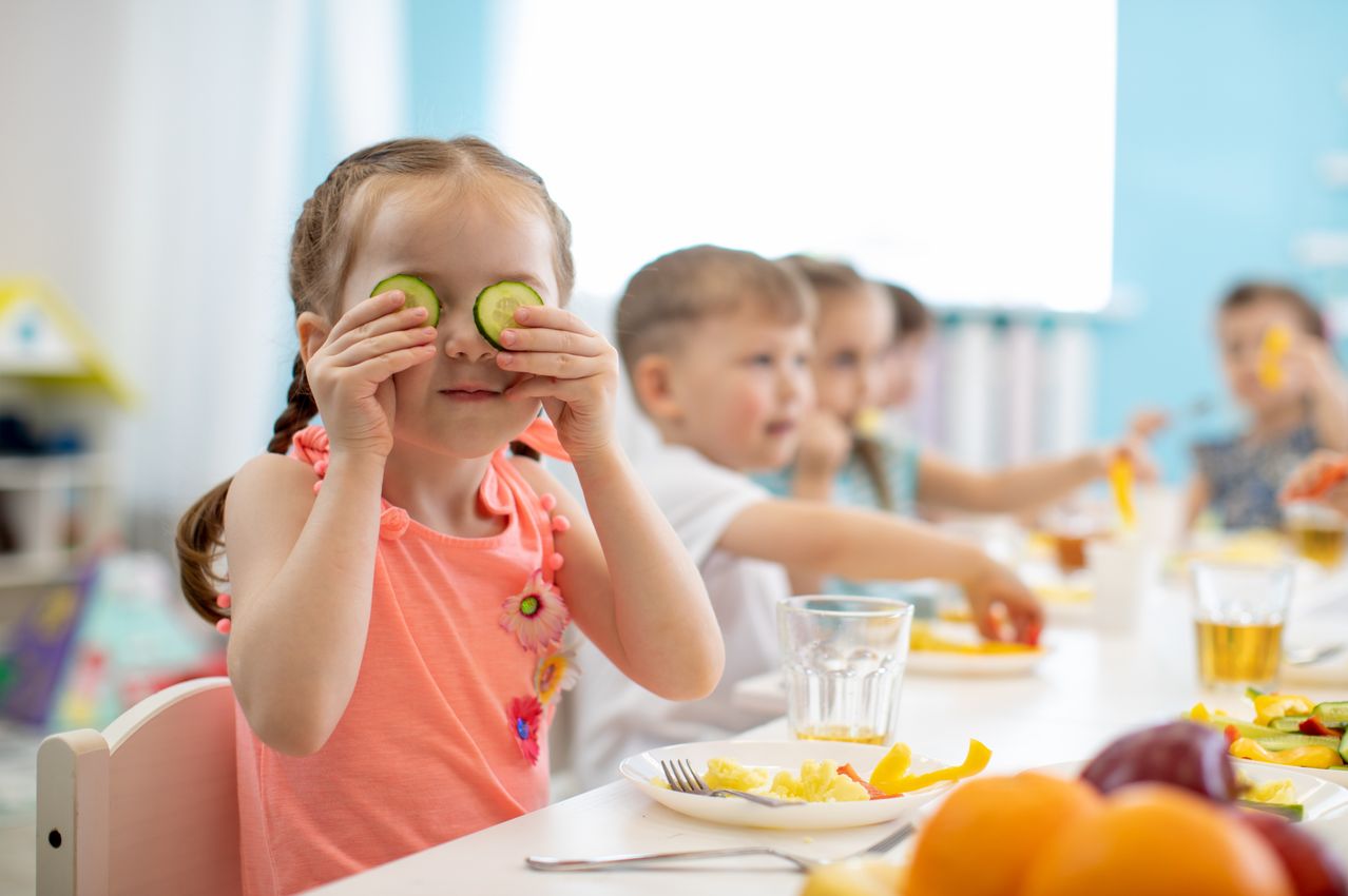 Ein kleines Mädchen an einem Tisch in einem Klassenzimmer, das Gurkenscheiben als Sonnenbrille benutzt. Hinter ihr sitzen weitere Kinder, die ebenfalls essen. Verschiedene Lebensmittel stehen auf dem Tisch, darunter Eier und ein Glas Saft.