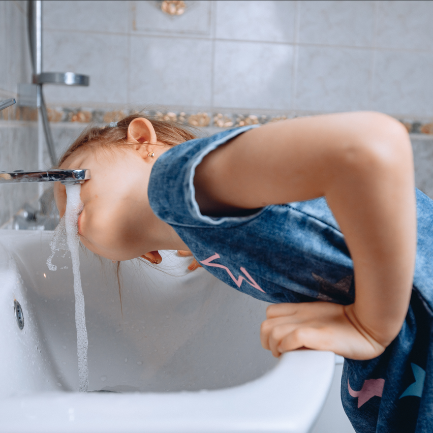 A young girl drinks water from the faucet in a bathroom. She leans forward over the sink with her mouth open.