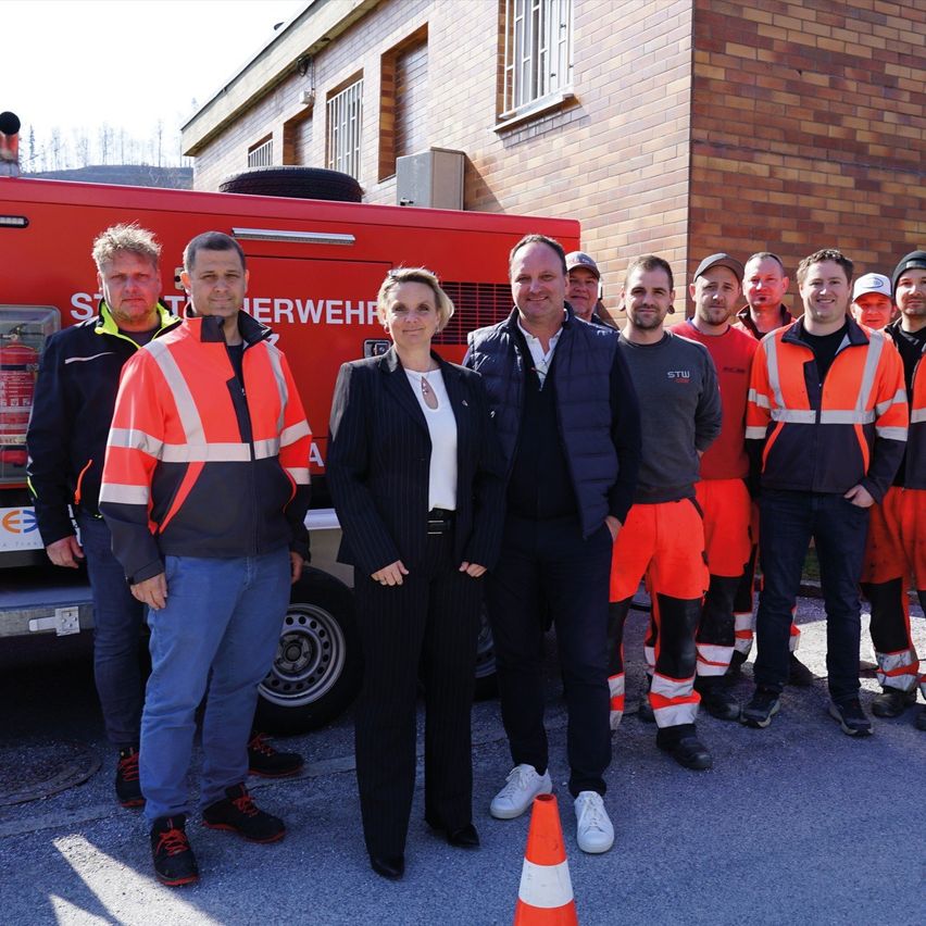 A group of individuals in high visibility safety suits stand in front of a fire truck. One woman stands out in formal attire.