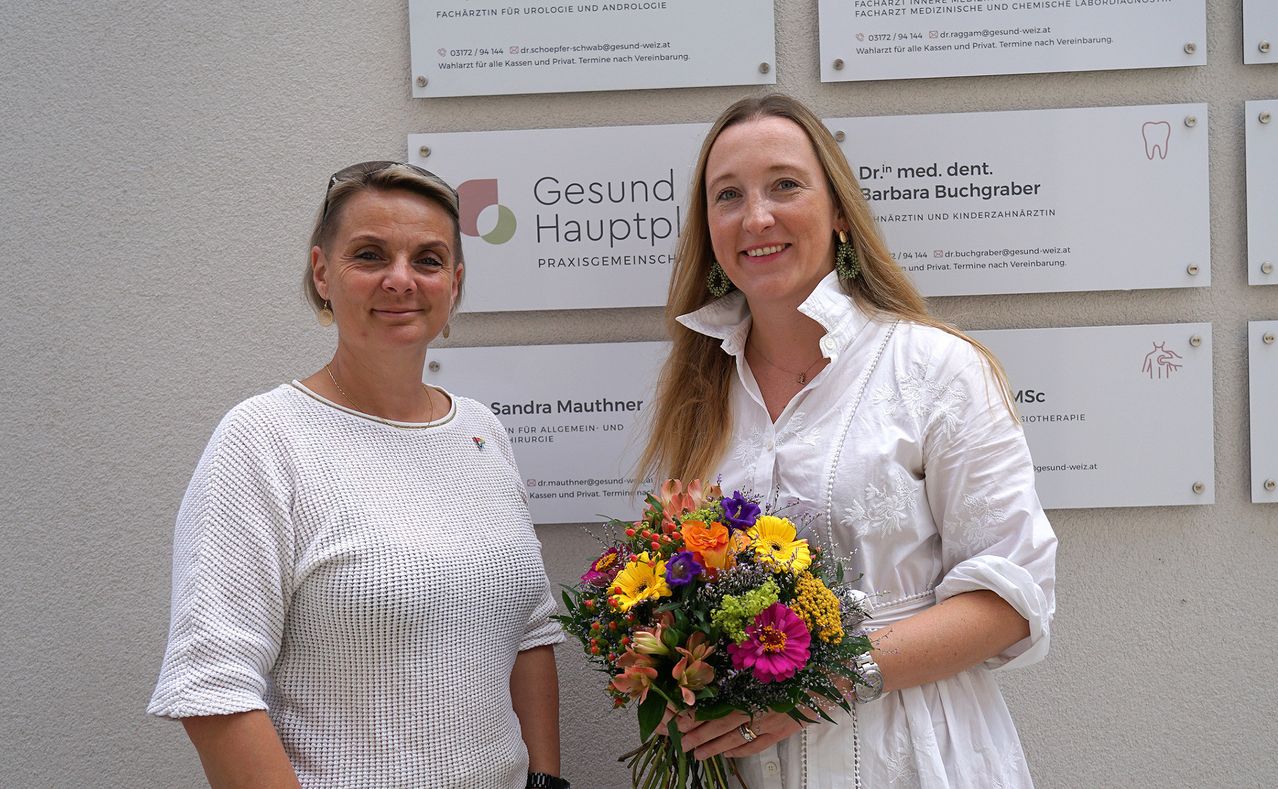 Two women stand smiling in front of a wall with medical practice signs. The woman on the left wears a white sweater, and the woman on the right holds a colorful bouquet.