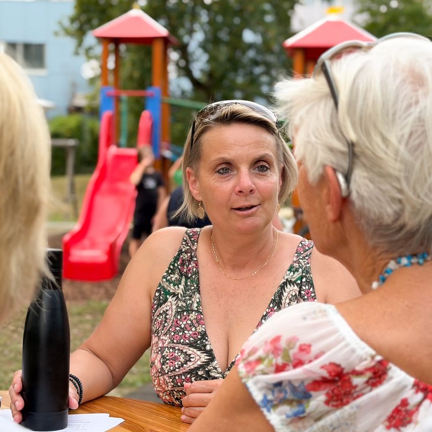 Eine Frau mit einer Brille auf dem Kopf sitzt an einem Tisch im Park und unterhält sich mit zwei anderen Frauen. Eine schwarze Flasche steht auf dem Tisch. Dahinter befindet sich ein Spielplatz mit einer roten Rutsche.