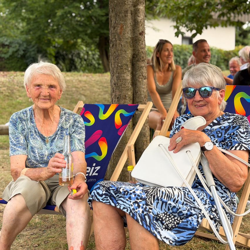 Zwei ältere Frauen sitzen in Liegestühlen unter einem Baum, eine hält eine Flasche und die andere hat eine Handtasche. Dahinter sitzen eine Frau und ein Mann auf einer Bank. Bäume und ein Gebäude sind im Hintergrund.