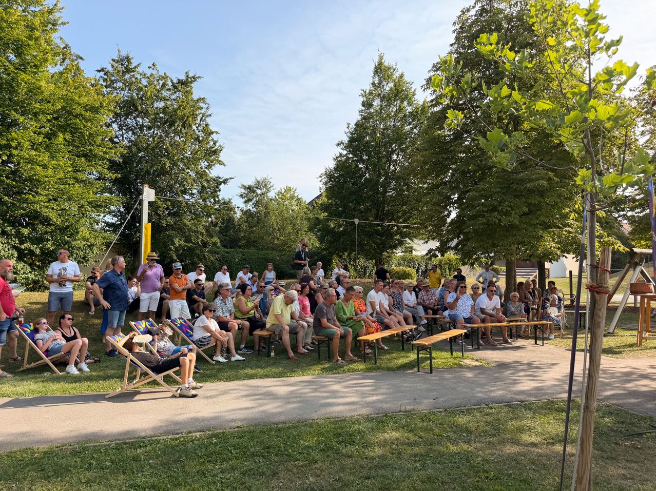 Eine Gruppe von Menschen sitzt auf Bänken in einem Park, einige stehen und reden. Bäume umgeben sie, und ein Pfahl mit einem gelben Schild steht links.