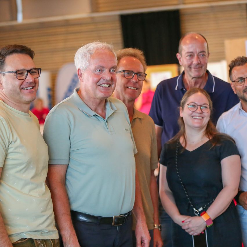 Eine Gruppe von sechs Personen, alle lächelnd, steht zusammen in einem gut beleuchteten Raum. Sie sind lässig gekleidet, einige tragen Brillen. Der Hintergrund ist unscharf, sodass der Fokus auf den Personen liegt.