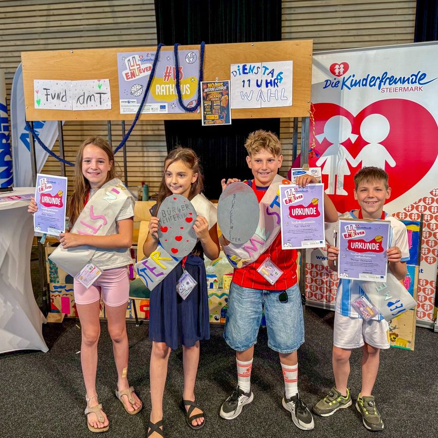 Fünf Kinder stehen zusammen und halten Urkunden in der Hand. Sie tragen Abzeichen und lächeln für ein Foto in einem Raum mit einem Hintergrund, der ein Herz und Kinderfiguren zeigt.