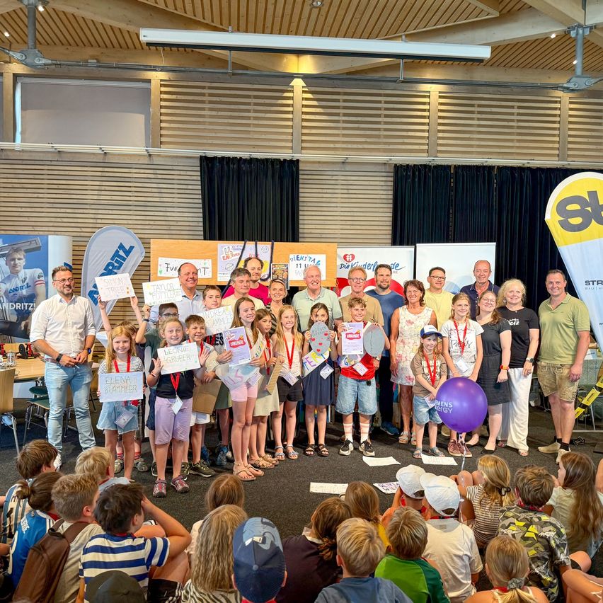 Eine Gruppe von Kindern und Erwachsenen steht auf einer Bühne, hält Urkunden und lächelt für ein Foto. Die Bühne ist mit Bannern und einem großen gelben Luftballon dekoriert.