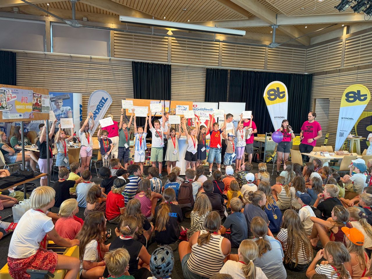 Eine Gruppe von Kindern und Erwachsenen versammelt sich in einem Raum, einige halten Schilder hoch, während andere sitzen. Eine Frau steht mit einem Mikrofon und einem Luftballon auf der Bühne.