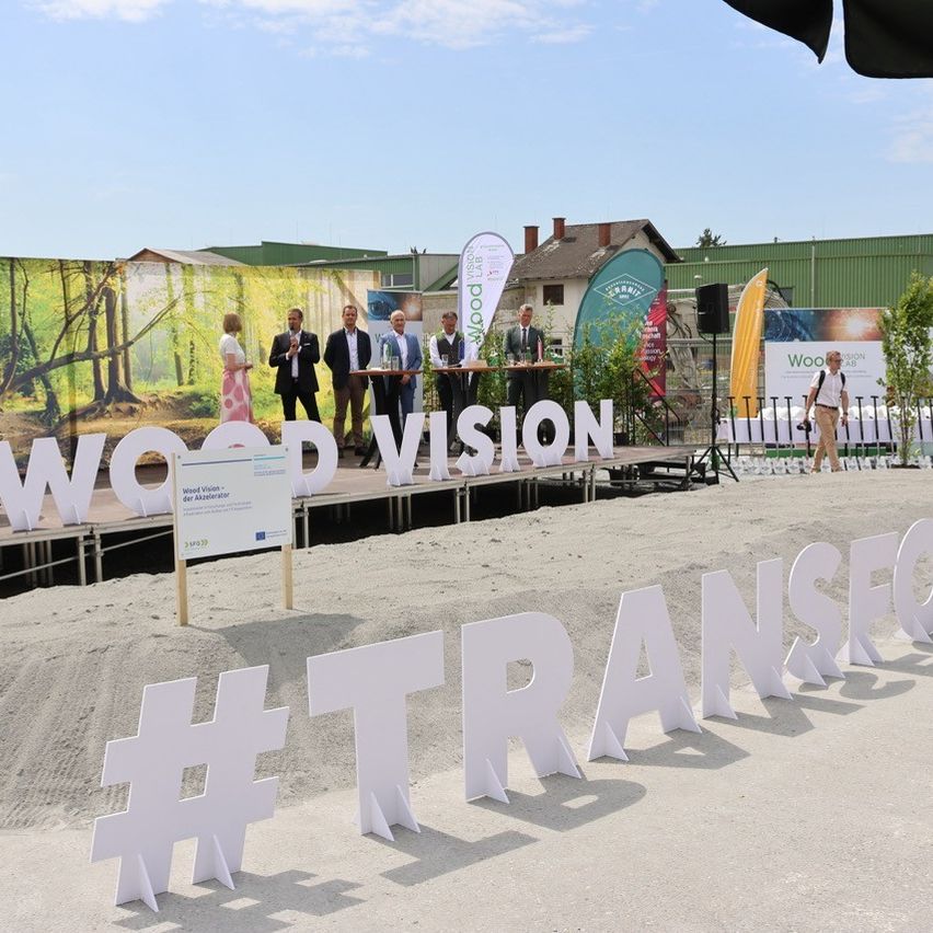 Eine Gruppe von Menschen steht auf einer Bühne mit einem Hintergrund, der 'Wood Vision' besagt. Sie sind umgeben von großen Buchstaben, die '#TRANSFO' auf dem Boden bilden. Eine Person geht im Hintergrund.