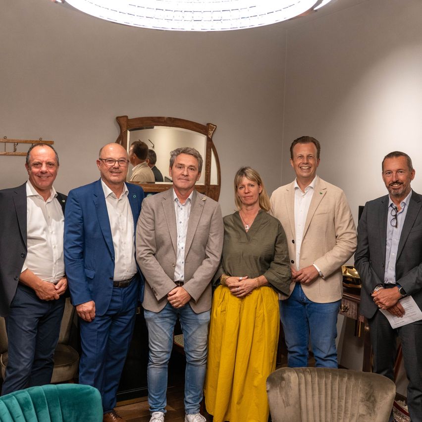Eine Gruppe von sechs Personen in formeller Kleidung steht lächelnd für ein Foto in einem Raum. Hinter ihnen befindet sich ein Spiegel mit hölzerne Rahmen und eine Lampe.