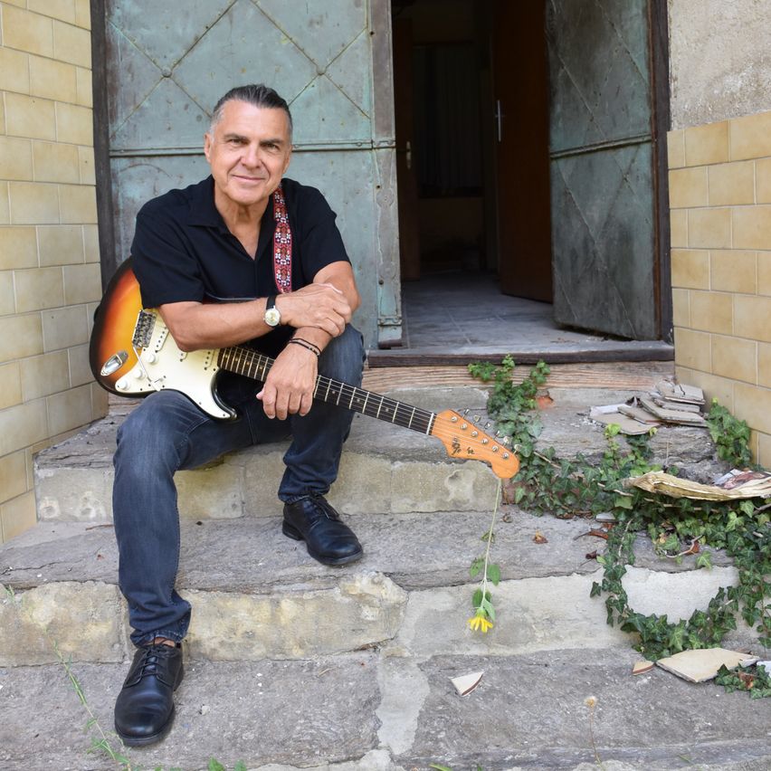 Ein Mann sitzt auf den Stufen eines Gebäudes, hält eine Gitarre und lächelt in die Kamera.