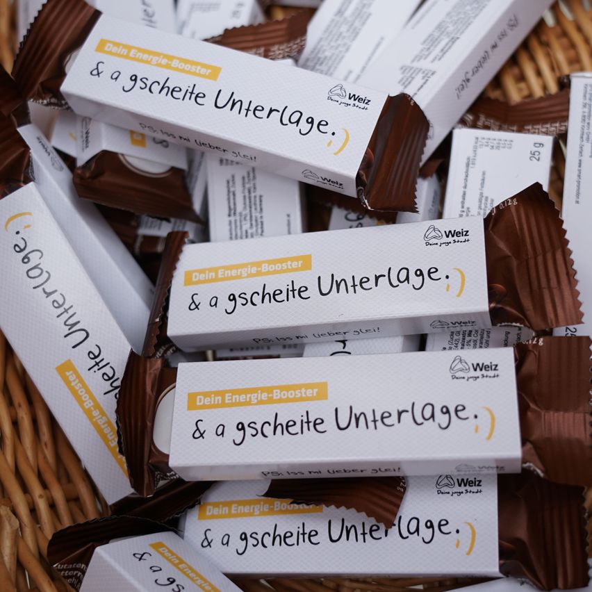 Ein Korb gefüllt mit mehreren Schokoladentafeln, die in weiße und gelbe Verpackungen eingewickelt sind, jede mit der Aufschrift 'Dein Energie-Booster & a schiefe Unterlage.' Die Schokoladentafeln befinden sich in verschiedenen Positionen, einige sind übereinander gestapelt.