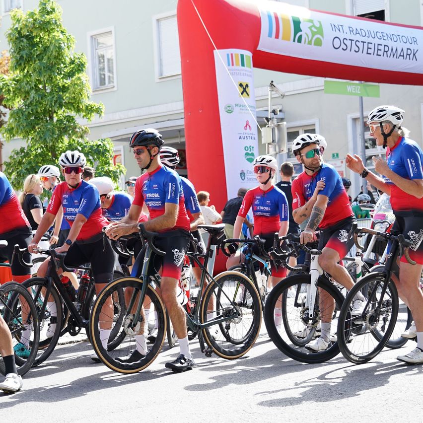 Eine Gruppe von Radfahrern in passenden blauen und roten Trikots steht in einer Reihe, möglicherweise zum Start eines Rennens. Sie stehen vor einem Banner und einem Gebäude.