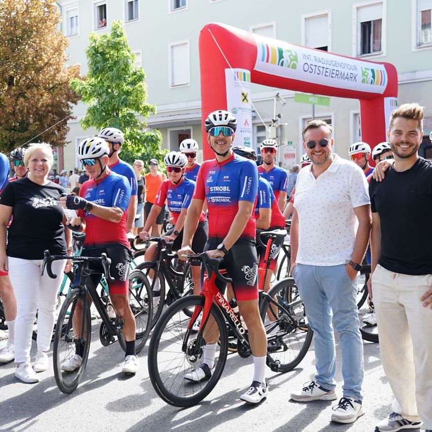 Eine Gruppe von Radfahrern in Radbekleidung, die neben einem roten Bogen mit dem Text 'Int. Radjugendtour Oststeiermark' stehen. Sie posieren für ein Foto mit zwei Männern in Freizeitkleidung.