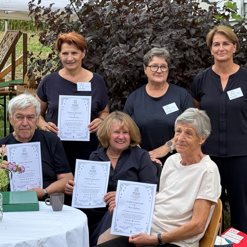 Eine Gruppe älterer Frauen und Männer posiert für ein Foto mit Zertifikaten in der Hand. Sie stehen vor einem Tisch mit einer Blumenvase und einer Tasse. Dahinter steht ein großer Busch.