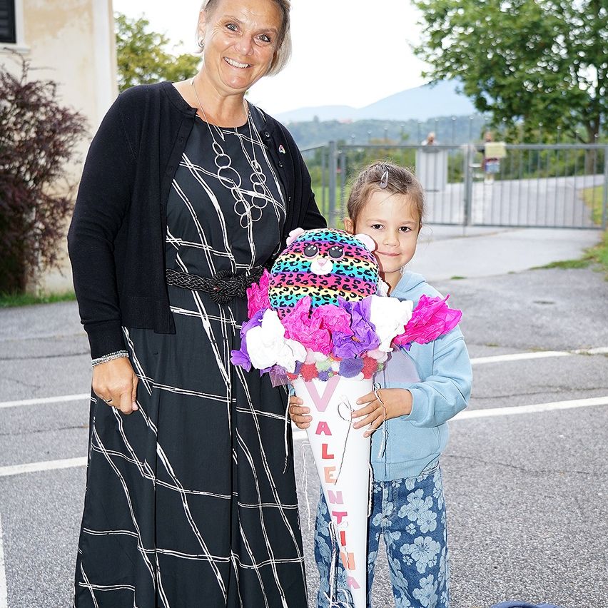 Eine Frau und ein kleines Mädchen stehen auf einem Parkplatz, das Mädchen hält einen Blumenstrauß in einem Kegel, lächelt und schaut zur Kamera.