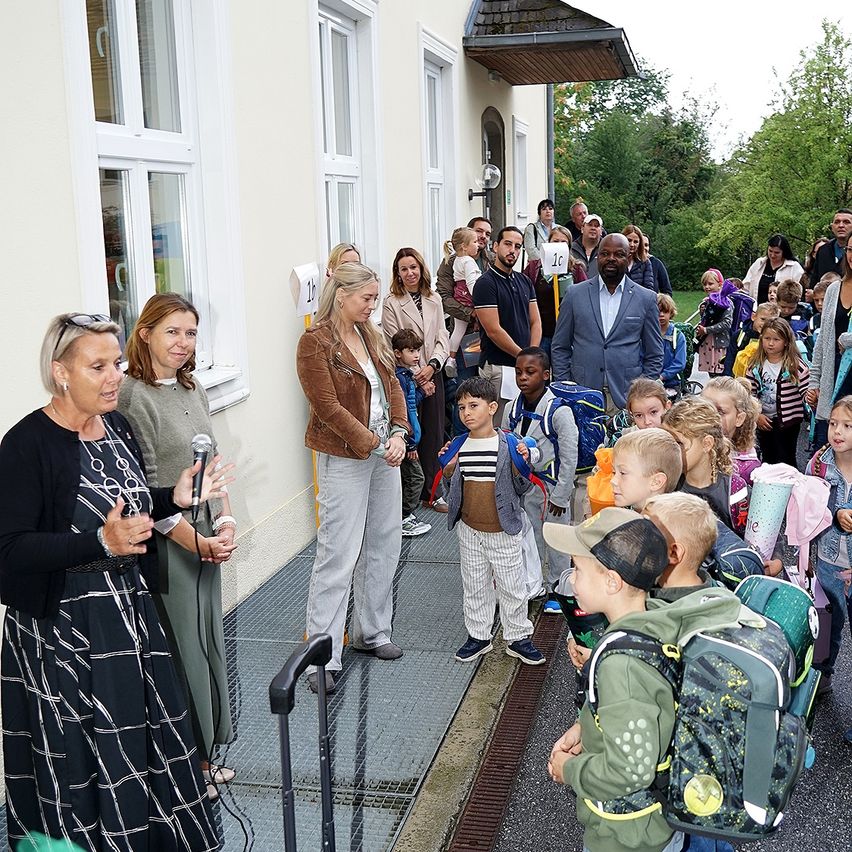 Eine Gruppe von Menschen, darunter Kinder, versammelt sich vor einem Gebäude. Eine Frau mit Mikrofon spricht zu ihnen. Die Kinder tragen Rucksäcke und Turnschuhe. Bäume und ein Haus sind im Hintergrund zu sehen.