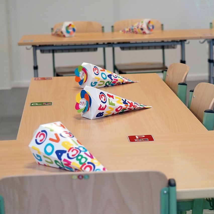 Ein Klassenzimmer mit Holztischen und Stühlen, jeweils mit bunten Kegeln darauf. Die Kegel sind mit Buchstaben und Zahlen verziert. Die Tische haben Etiketten und ein rotes Schild. Der Raum ist gut beleuchtet.