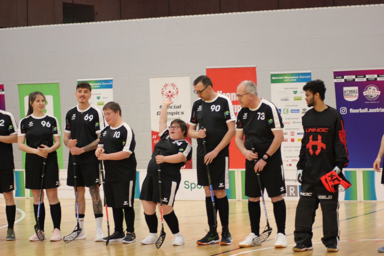 Eine Gruppe von Personen in Sportuniformen steht in einer Reihe und hält Lacrosse-Schläger. Sie scheinen sich auf ein Spiel vorzubereiten. Der Ort ist drinnen, mit Bannern an der Wand hinter ihnen.