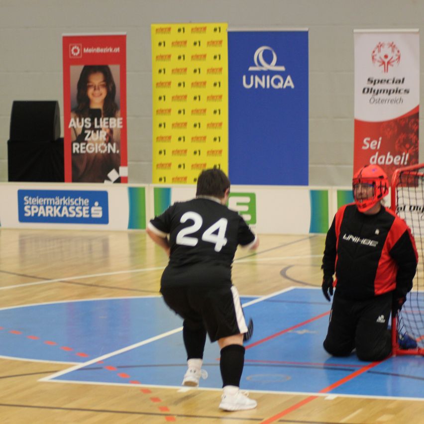 Zwei Spieler spielen Futsal auf einem Hallenspielfeld. Ein Spieler trägt einen roten Helm und kniet vor dem Tor. Der andere Spieler trägt ein schwarzes Trikot mit der Nummer 24.