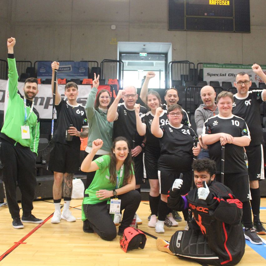 Eine Gruppe von Personen, wahrscheinlich ein Sportteam, posiert für ein Foto in einer Indoor-Sportanlage. Sie tragen alle einheitliche Kleidung und einige halten Hockeyschläger.