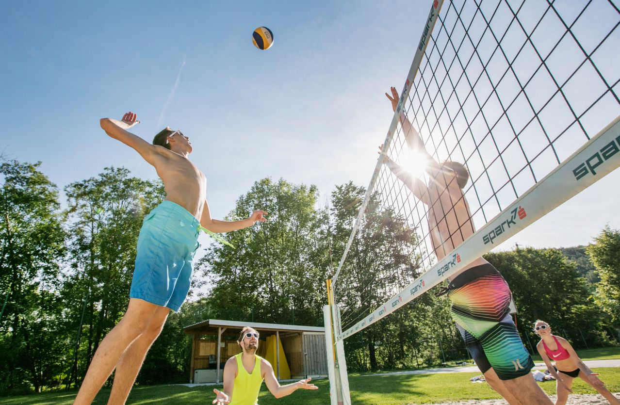 Drei Männer spielen Beachvolleyball in einem Park, einer greift nach oben, um den Ball zu blockieren. Ein anderer Spieler springt, um den Ball zu spiken.